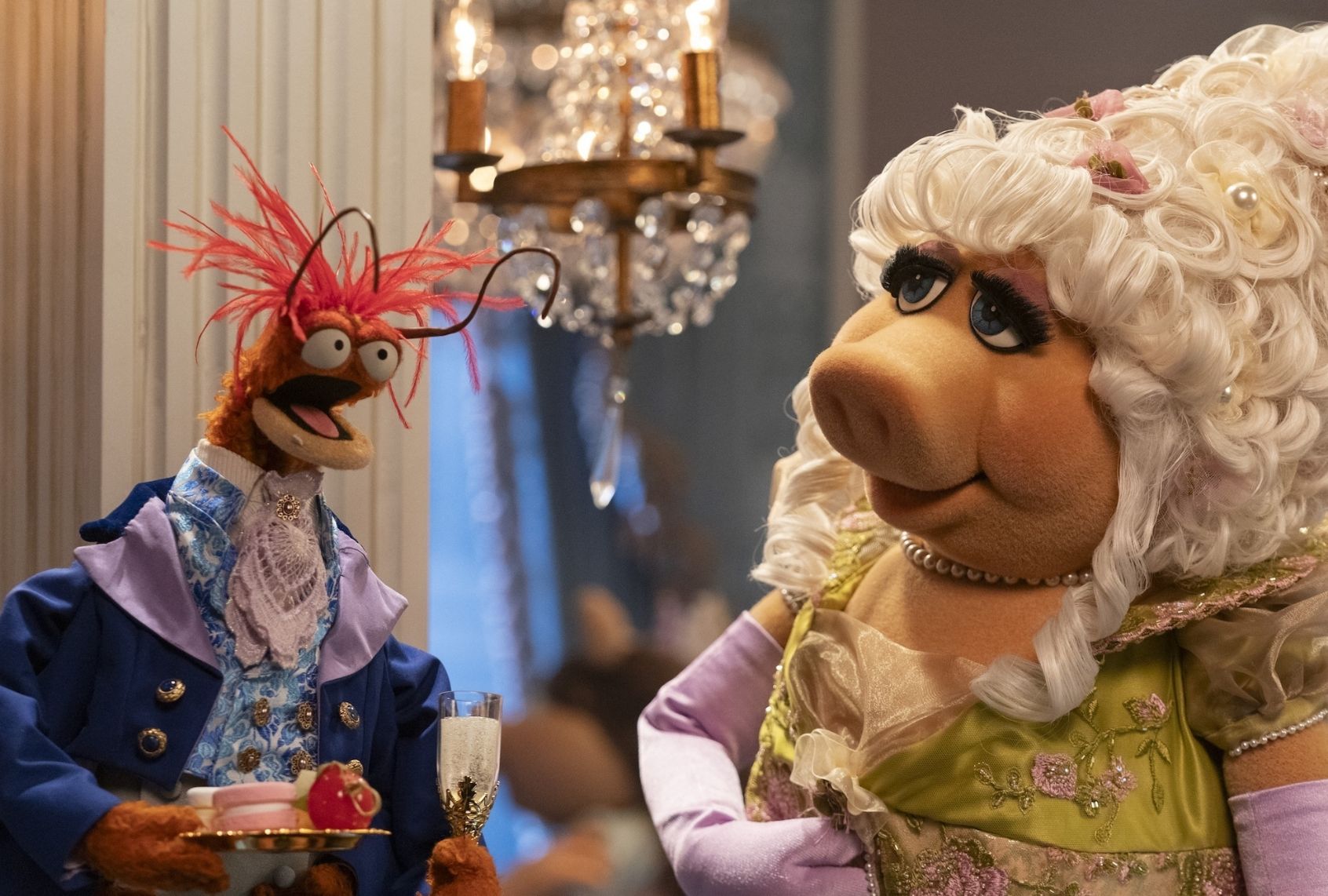 A pig puppet with blonde hair styled into a curly updo with face-framing tendrils is attired for a ball in a chartreuse and pink puffl-sleeved gown, lavender elbow gloves and a string of pearls. She is taking to a raffish orange prawn with red hair wearing a navy suit jacket over a cornflower blue-patterned vest and white lace cravat. He is holding a goblet and a plate full of macarons and other sweets. They're in a ballroom with a crystal chandelier